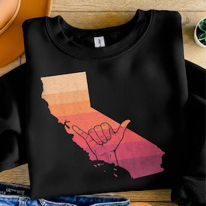May include: Black sweatshirt featuring a graphic of the California state outline with a sunset gradient and a hand making the shaka sign. The design is in shades of orange, red, and pink.