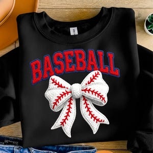 May include: Black sweatshirt with the word "BASEBALL" in red, outlined in blue, above a white bow with red baseball stitching. The bow is the focal point, with detailed stitching.