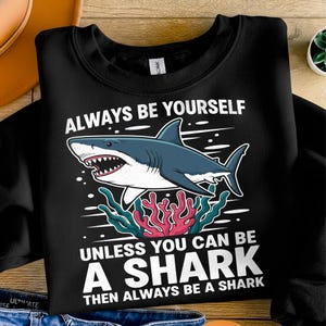 Könnte beinhalten: Schwarzes Sweatshirt mit einem Hai-Motiv und dem Text "ALWAYS BE YOURSELF UNLESS YOU CAN BE A SHARK THEN ALWAYS BE A SHARK". Der Hai ist blau und weiß, mit roter und grüner Koralle.