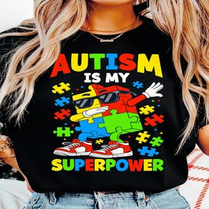 Puede incluir: Camiseta negra con el texto "AUTISM IS MY SUPERPOWER" en letras coloridas. El diseño presenta piezas de rompecabezas en rojo, amarillo, azul y verde, con un personaje de pieza de rompecabezas con gafas de sol y zapatillas rojas.