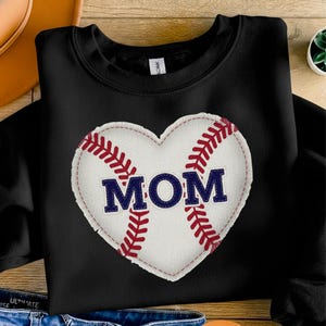 May include: Black sweatshirt featuring a white heart-shaped baseball with red stitching and the word "MOM" in navy blue. The design is centered on the front of the sweatshirt, making it a perfect gift for baseball moms.