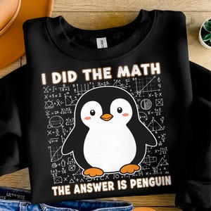 Op de afbeelding: Zwarte sweatshirt met de tekst "I DID THE MATH THE ANSWER IS PENGUIN" en een cartoon pinguïn. De pinguïn is zwart en wit met oranje voeten en snavel. Wiskundige vergelijkingen op de achtergrond.