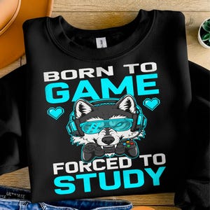Może przedstawiać: Czarna bluza z napisem "BORN TO GAME FORCED TO STUDY" w kolorze białym i turkusowym. Kreskówkowy wilk w turkusowych okularach i słuchawkach trzyma kontroler do gier. Dwa turkusowe pikselowe serca znajdują się po obu stronach wilka.