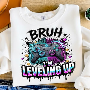 Puede incluir: Sudadera blanca con un gráfico de un mando de videojuegos. El mando es negro, turquesa y morado, con las palabras "BRUH I'M LEVELING UP" en negro, turquesa y morado. El diseño incluye estrellas, corazones y monedas.