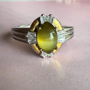 May include: A silver ring with a gold and white accent. The ring features a large, oval, olive-green gemstone at the center, surrounded by a gold setting and white accent stones. The band has a ribbed design. The ring is displayed on a pink surface.
