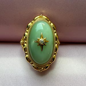 May include: A gold-toned ring with a light green oval stone. A starburst design with a small clear stone is set in the center of the oval. The ring has a decorative, ornate border.
