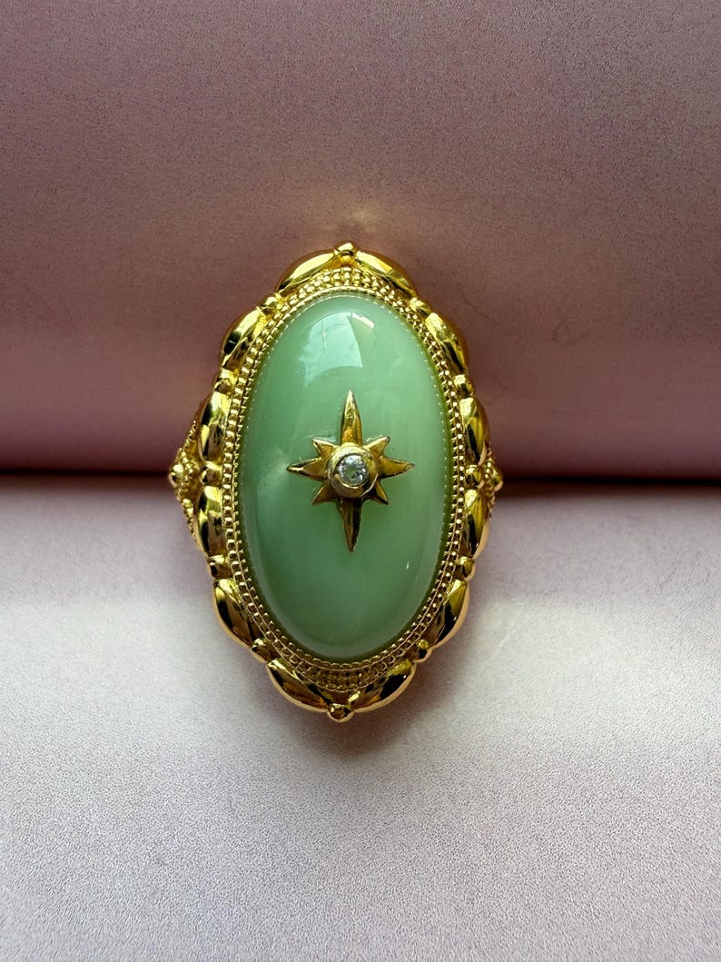 K&ouml;nnte beinhalten: Ein goldfarbener Ring mit einem hellgr&uuml;nen ovalen Stein. Ein Sternmuster mit einem kleinen, klaren Stein befindet sich in der Mitte des Ovals. Der Ring hat einen dekorativen, verzierten Rand.