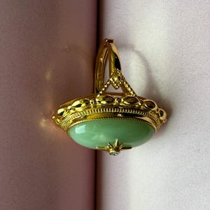 K&ouml;nnte beinhalten: Ein goldfarbener Ring mit einem gro&szlig;en, ovalen, hellgr&uuml;nen Stein. Der Ring hat aufwendige Details um den Stein herum und einen kleinen, klaren Akzentstein. Der Ring ist vor einem rosa Hintergrund platziert.