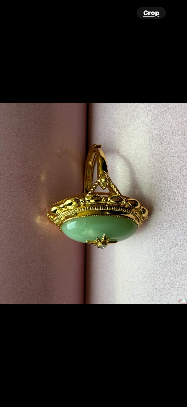 K&ouml;nnte beinhalten: Ein goldfarbener Ring mit einem gro&szlig;en, ovalen, hellgr&uuml;nen Stein. Der Ring hat aufwendige Details um den Stein herum und einen kleinen, klaren Akzentstein. Der Ring ist vor einem rosa Hintergrund platziert.