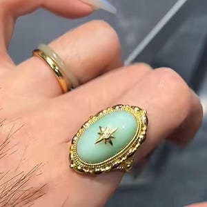 K&ouml;nnte beinhalten: Goldener Ring mit einem hellgr&uuml;nen, ovalen Stein und einem Sternmuster. Der Ring hat einen detaillierten goldenen Filigranrand. Ein zweiter goldener Ring mit einem hellgr&uuml;nen Band ist ebenfalls sichtbar.