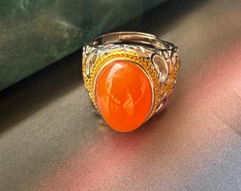 Könnte beinhalten: Ein silberner und goldener Ring mit einem großen, ovalen, orangefarbenen Edelstein. Der Ring hat aufwendige Filigranarbeiten um den Edelstein und entlang des Rings. Der Edelstein ist der Mittelpunkt mit einer polierten, reflektierenden Oberfläche.