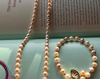 Könnte beinhalten: Ein Halsketten- und Armband-Set mit ovalen Perlen in Pfirsich-, Creme- und Grautönen. Die Halskette ist lang, während das Armband einen goldfarbenen Anhänger mit einem Perlenanhänger hat. Das Schmuckset liegt auf einer Magazinseite.