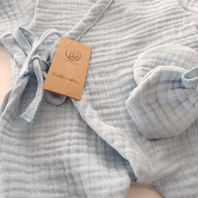 K&ouml;nnte beinhalten: Hellblaues Baby-Set aus weichem, strukturiertem Stoff, bestehend aus einem Wickelbody und passenden F&auml;ustlingen. Der Body hat ein Wickeldesign mit Bindeb&auml;ndern. Ein braunes Etikett mit Logo und Text ist angebracht.