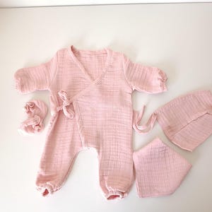 K&ouml;nnte beinhalten: Ein hellrosa Baby-Kleidungsset, bestehend aus einem Wickel-Overall, Booties, einem L&auml;tzchen und einer M&uuml;tze. Das Set ist aus einem weichen, strukturierten Stoff gefertigt. Der Overall hat lange &Auml;rmel und Bindeb&auml;nder an der Taille.