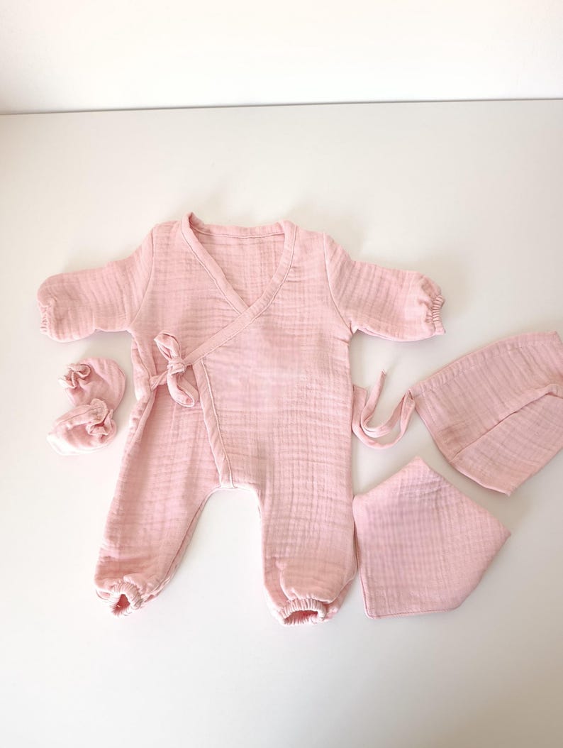 K&ouml;nnte beinhalten: Ein hellrosa Baby-Kleidungsset, bestehend aus einem Wickel-Overall, Booties, einem L&auml;tzchen und einer M&uuml;tze. Das Set ist aus einem weichen, strukturierten Stoff gefertigt. Der Overall hat lange &Auml;rmel und Bindeb&auml;nder an der Taille.