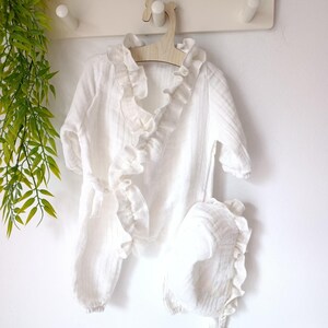 May include: A white long-sleeved wrap-style romper featuring a ruffled trim along the neckline and front opening, accompanied by a matching bonnet with a ruffled edge. The garment is made of a textured fabric and includes a side tie closure.