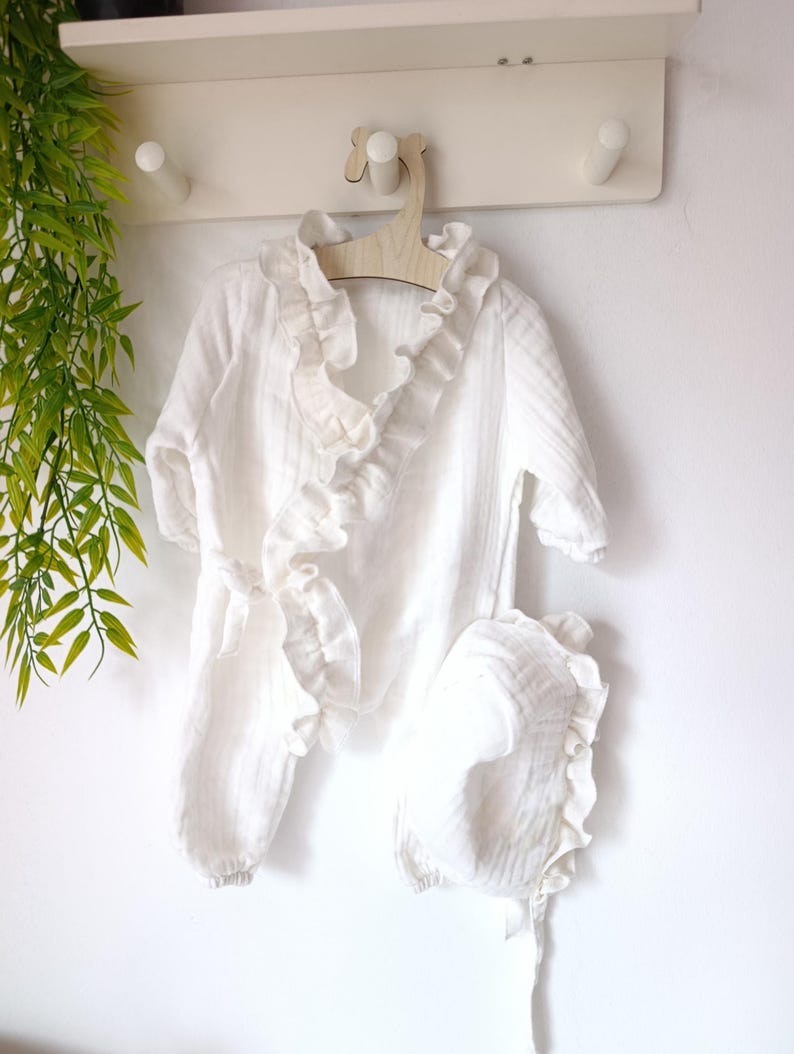 May include: A white long-sleeved wrap-style romper featuring a ruffled trim along the neckline and front opening, accompanied by a matching bonnet with a ruffled edge. The garment is made of a textured fabric and includes a side tie closure.