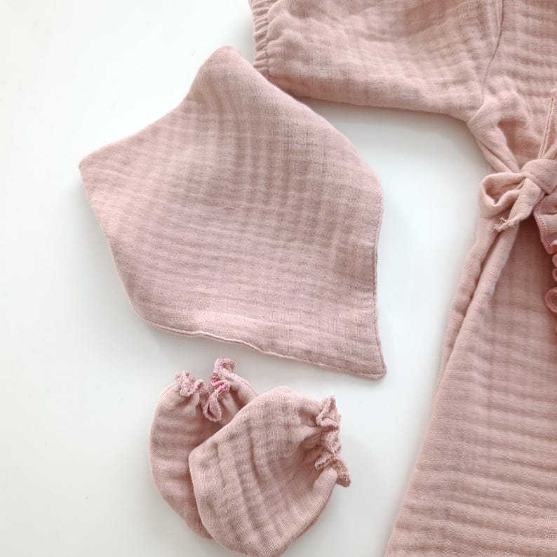 May include: A dusty pink muslin set consisting of a long-sleeved garment with a side tie closure, a triangular bib, and a pair of small mittens. The fabric features a soft, crinkled texture throughout all pieces, showcasing a consistent light rose color.