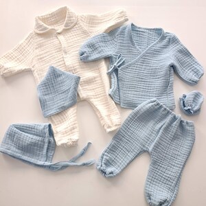 K&ouml;nnte beinhalten: Eine Kollektion von Babykleidung aus weichem, strukturiertem Stoff. Das Set beinhaltet einen cremefarbenen Overall, ein hellblaues Wickeloberteil, eine Hose, ein L&auml;tzchen und eine M&uuml;tze. Die Kleidungsst&uuml;cke sind aus leichtem Material gefertigt.
