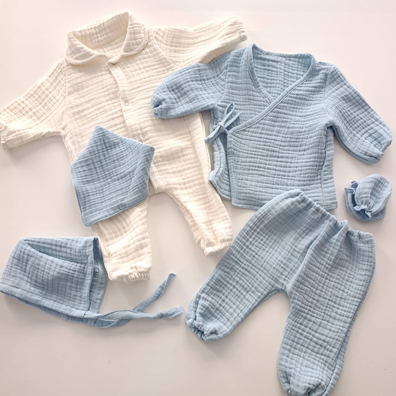 K&ouml;nnte beinhalten: Eine Kollektion von Babykleidung aus weichem, strukturiertem Stoff. Das Set beinhaltet einen cremefarbenen Overall, ein hellblaues Wickeloberteil, eine Hose, ein L&auml;tzchen und eine M&uuml;tze. Die Kleidungsst&uuml;cke sind aus leichtem Material gefertigt.