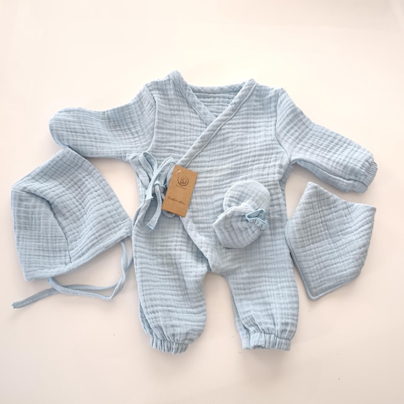 K&ouml;nnte beinhalten: Hellblaues Baby-Outfit-Set, bestehend aus einem Wickel-Jumpsuit mit langen &Auml;rmeln, einer M&uuml;tze, F&auml;ustlingen und einem L&auml;tzchen. Das Set ist aus einem weichen, strukturierten Stoff gefertigt. Ein braunes Etikett ist am Jumpsuit befestigt.