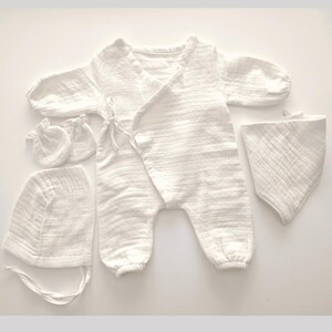 K&ouml;nnte beinhalten: Ein wei&szlig;es Baby-Kleidungsset, bestehend aus einem Wickel-Overall, einer M&uuml;tze, einem L&auml;tzchen und einem Paar F&auml;ustlinge. Das Set ist aus einem weichen, strukturierten Stoff gefertigt. Der Overall hat lange &Auml;rmel und Bindeb&auml;nder an der Taille.