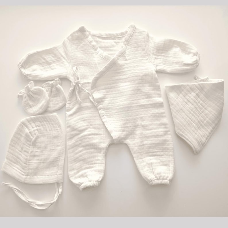 K&ouml;nnte beinhalten: Ein wei&szlig;es Baby-Kleidungsset, bestehend aus einem Wickel-Overall, einer M&uuml;tze, einem L&auml;tzchen und einem Paar F&auml;ustlinge. Das Set ist aus einem weichen, strukturierten Stoff gefertigt. Der Overall hat lange &Auml;rmel und Bindeb&auml;nder an der Taille.