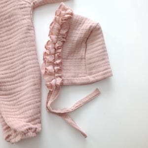 May include: A dusty pink muslin bonnet featuring a ruffled trim along the front edge and two fabric ties for fastening, positioned next to a matching long-sleeved garment made of the same textured, lightweight, crinkled fabric material.