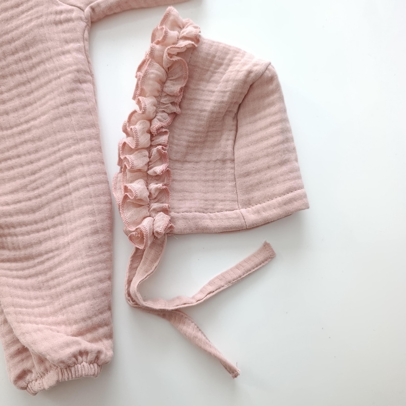 May include: A dusty pink muslin bonnet featuring a ruffled trim along the front edge and two fabric ties for fastening, positioned next to a matching long-sleeved garment made of the same textured, lightweight, crinkled fabric material.