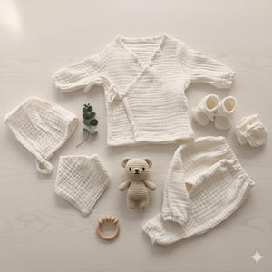 K&ouml;nnte beinhalten: Eine Auswahl an Babykleidung und Accessoires in Creme. Das Set beinhaltet ein Wickeloberteil, eine Hose, eine M&uuml;tze, ein L&auml;tzchen, Babyschuhe und einen kleinen geh&auml;kelten Teddyb&auml;ren. Ein Bei&szlig;ring aus Holz und ein Zweig Gr&uuml;nzeug sind ebenfalls enthalten.