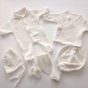 K&ouml;nnte beinhalten: Ein Set cremefarbener Babykleidung, darunter ein Strampler mit Kragen, ein Wickeloberteil, eine Hose, eine M&uuml;tze, ein L&auml;tzchen und F&auml;ustlinge. Die Artikel sind aus einem weichen, strukturierten Stoff gefertigt.