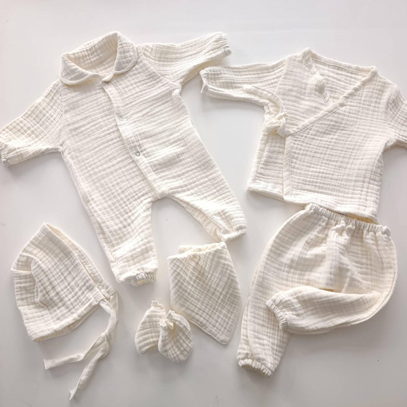 K&ouml;nnte beinhalten: Ein Set cremefarbener Babykleidung, darunter ein Strampler mit Kragen, ein Wickeloberteil, eine Hose, eine M&uuml;tze, ein L&auml;tzchen und F&auml;ustlinge. Die Artikel sind aus einem weichen, strukturierten Stoff gefertigt.