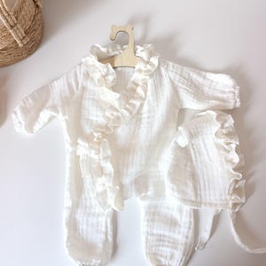 May include: A white long-sleeved romper with a ruffled collar and front trim, featuring elasticized cuffs at the wrists and ankles. The set includes a matching white bonnet with a ruffled edge and tie closures, all made from a textured fabric.
