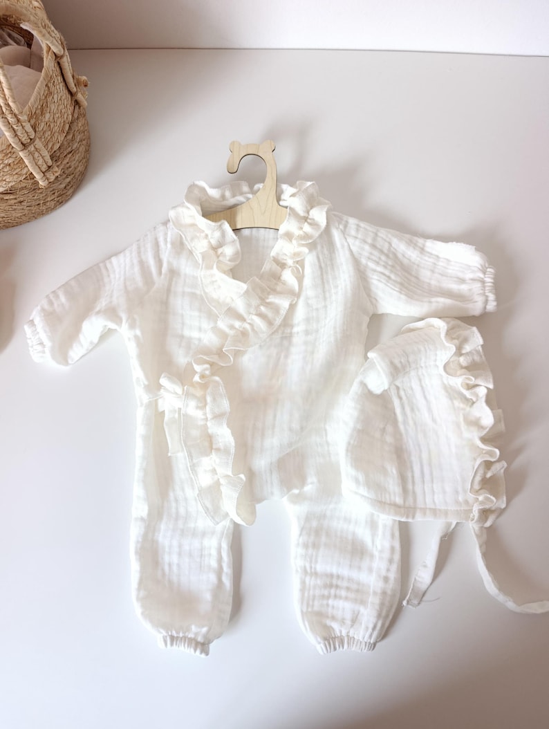 May include: A white long-sleeved romper with a ruffled collar and front trim, featuring elasticized cuffs at the wrists and ankles. The set includes a matching white bonnet with a ruffled edge and tie closures, all made from a textured fabric.