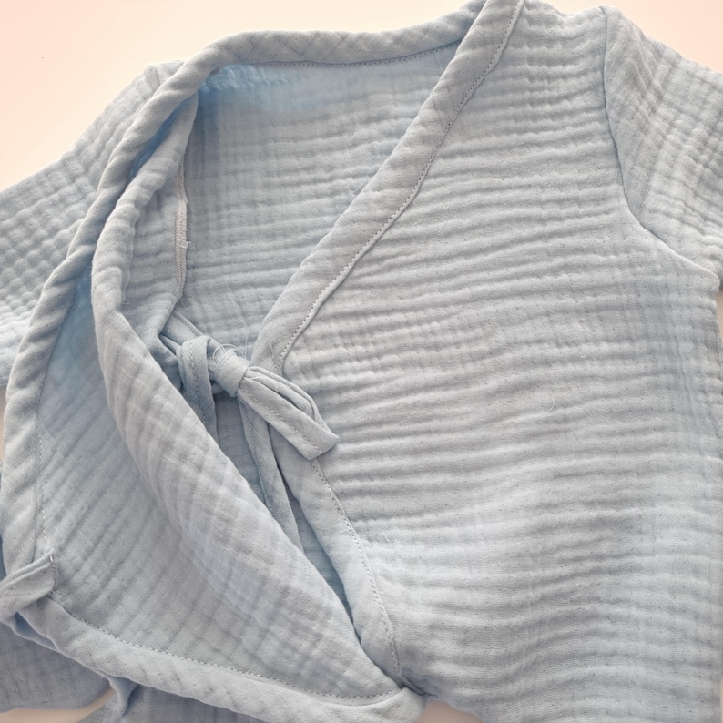 K&ouml;nnte beinhalten: Hellblaue Babyrobe aus weichem, strukturiertem Stoff. Die Robe hat ein Wickeldesign mit einem Bindeband in der Taille. Die &Auml;rmel sind kurz und die Kanten sind mit passendem Besatz versehen. Die Robe ist auf Komfort und einfache Handhabung ausgelegt.