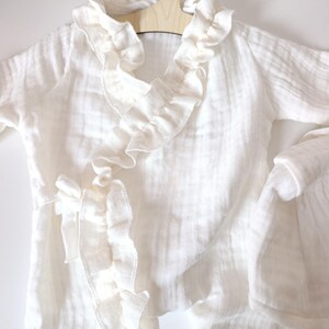 May include: A white long-sleeved wrap-style garment made of textured muslin fabric. It features a ruffled trim along the V-neckline and front opening, with a fabric tie closure at the side. The piece is displayed on a light-colored wooden hanger.
