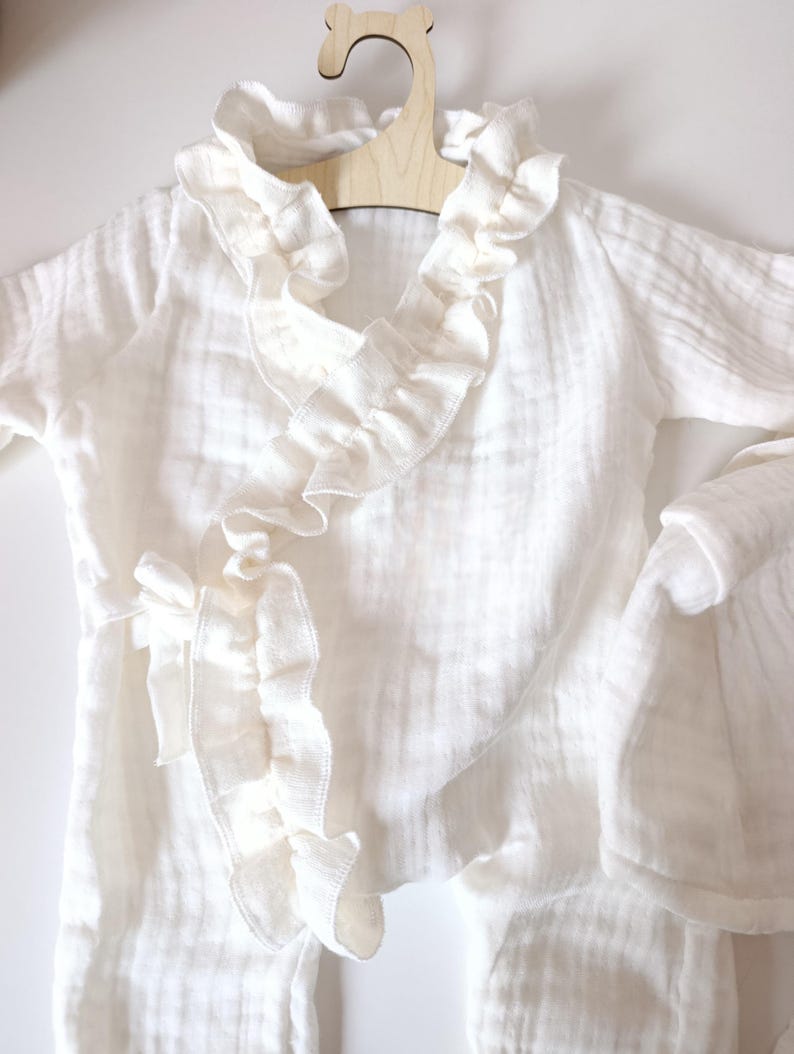 May include: A white long-sleeved wrap-style garment made of textured muslin fabric. It features a ruffled trim along the V-neckline and front opening, with a fabric tie closure at the side. The piece is displayed on a light-colored wooden hanger.
