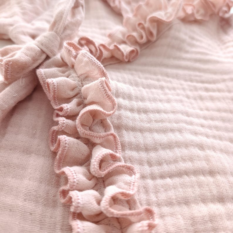 May include: A light pink muslin fabric garment featuring a textured, crinkled surface and delicate ruffled trim along the edges. The piece includes a fabric tie closure, showcasing the soft, layered folds of the material and the stitched detailing.