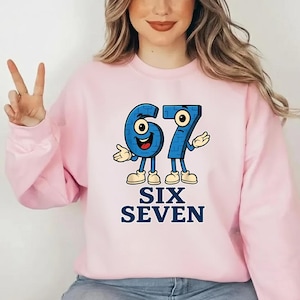 May include: A light pink sweatshirt featuring a cartoon design of the numbers 67 in blue, with arms, legs, and smiling faces. The words "SIX SEVEN" are printed below in navy blue.