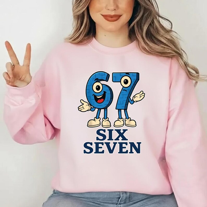 May include: A light pink sweatshirt featuring a cartoon design of the numbers 67 in blue, with arms, legs, and smiling faces. The words "SIX SEVEN" are printed below in navy blue.
