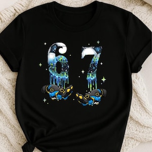 May include: Black t-shirt featuring the number "67" in a space-themed design with dripping blue and green accents. The numbers appear to be held by two gloved hands with blue and yellow details, surrounded by stars.