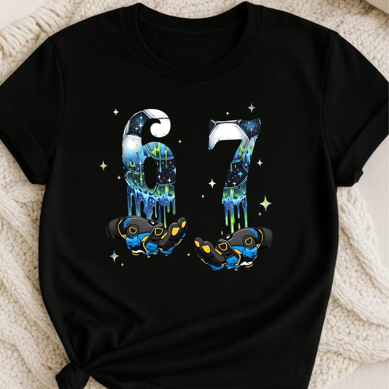 May include: Black t-shirt featuring the number "67" in a space-themed design with dripping blue and green accents. The numbers appear to be held by two gloved hands with blue and yellow details, surrounded by stars.