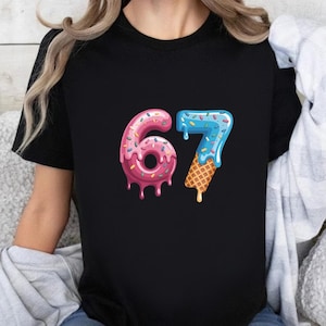 May include: Black t-shirt featuring the number 67 in a playful, dessert-themed design. The number 6 is pink with sprinkles and dripping icing, while the 7 is blue with a waffle cone base and dripping icing.