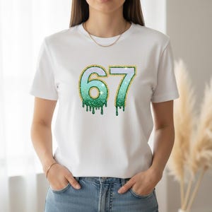 May include: White t-shirt featuring the number "67" in a teal and green glitter design with gold glitter trim. The numbers have a dripping effect. The shirt is worn by a person in blue jeans.