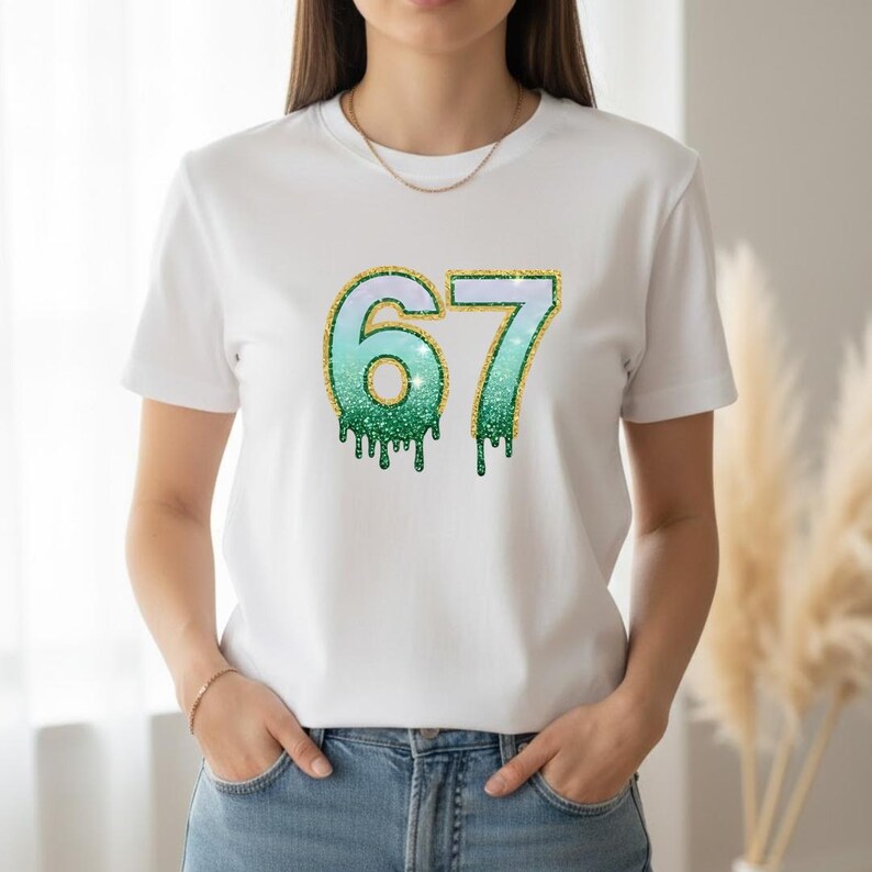 May include: White t-shirt featuring the number "67" in a teal and green glitter design with gold glitter trim. The numbers have a dripping effect. The shirt is worn by a person in blue jeans.