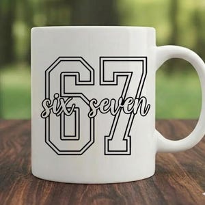 Può includere: Tazza in ceramica bianca con il numero 67 in contorno nero. Le parole "six seven" sono scritte in corsivo all'interno dei numeri. La tazza ha un manico curvo ed è appoggiata su una superficie di legno.