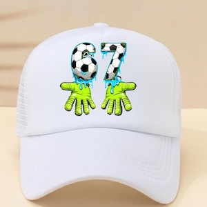 May include: White baseball cap with a mesh back and a curved brim. The cap features a graphic of the numbers "67" in a soccer ball design, with green hands and blue dripping accents. The cap is a sporty accessory.