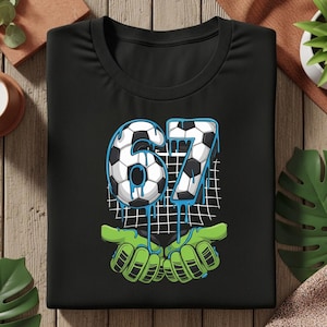 May include: Black t-shirt featuring a soccer-themed graphic. The design includes the number "67" stylized as soccer balls with blue dripping accents, set against a net, and held by green goalie gloves.