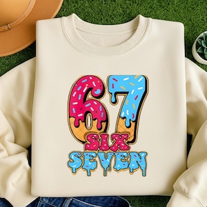May include: Cream-colored sweatshirt featuring a colorful graphic of the number "67" designed to look like a donut and ice cream cone. The words "SIX SEVEN" are below the numbers in a matching font.