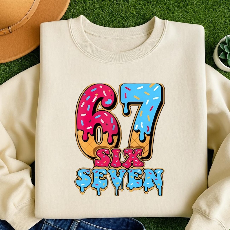 May include: Cream-colored sweatshirt featuring a colorful graphic of the number "67" designed to look like a donut and ice cream cone. The words "SIX SEVEN" are below the numbers in a matching font.